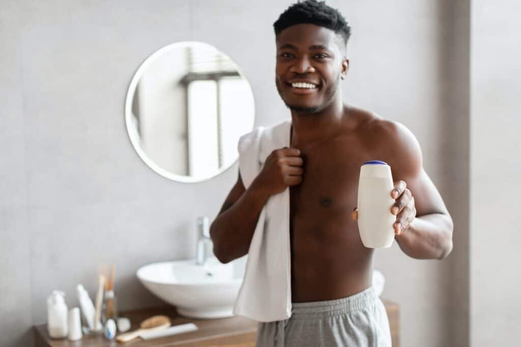 African Guy Showing Shampo Bottle Advertising Male Cosmetic In Bathroom
